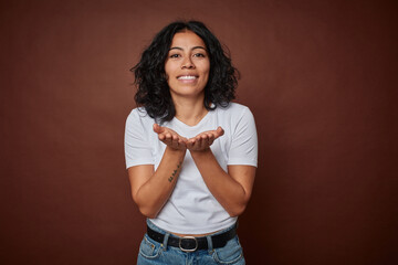 Young colombian curly hair woman isolated on brown background holding something with palms, offering to camera.