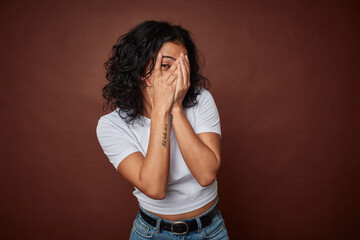 Young colombian curly hair woman isolated on brown background blink through fingers frightened and...