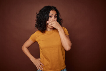 Young colombian curly hair woman isolated on brown background covering mouth with hands looking...