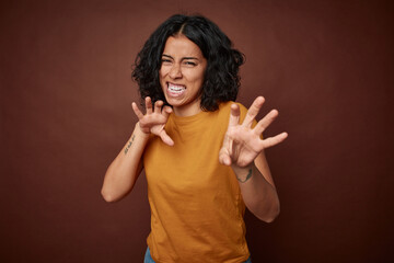 Young colombian curly hair woman isolated on brown background showing claws imitating a cat,...