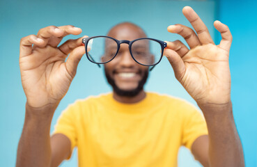 Fototapeta premium Hands, glasses and vision with a black man in studio on a blue background for prescription frame lenses. Spectacles, eyesight and eyewear with a male indoor to promote new round frames for seeing