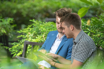Two businessman or gay sitting at chair in park while using digital tablet and paper work plan for...