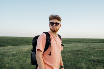 A bearded young man in sunglasses and a pink t-shirt stands on an endless Ukrainian field at sunset and looks away.