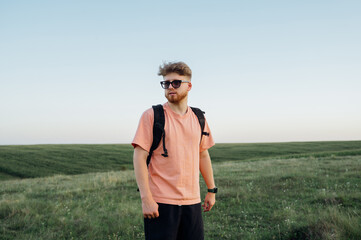 Positive bearded young man in casual clothes with a backpack on his back in the evening at sunset posing for the camera with a serious face and looking away.