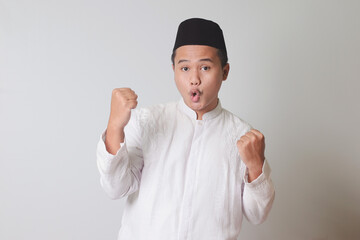 Portrait of Asian muslim man in white koko shirt with skullcap raising his fist, celebrating...