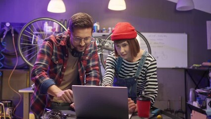 Young attractive professional worker of modern workshop buying parts for bike online using laptop with handsome professional craftsman. Cute lovely couple having family business.