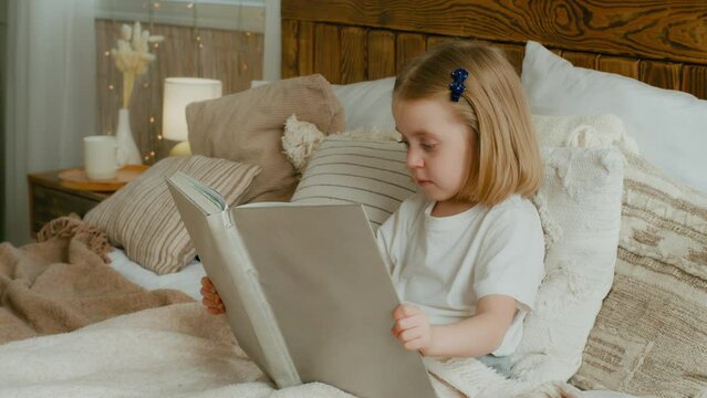 Cute little 5s Caucasian alone child girl daughter looking in book kid preschool baby sitting in bed watching pictures in copybook studying learning read try reading elementary education in bedroom