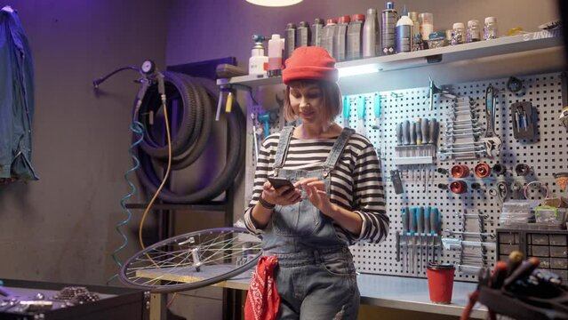 Portrait of attractive young caucasian woman standing at modern bike workshop using and typing on smartphone online smiling. Beautiful skillful female mechanic having break after hard work.