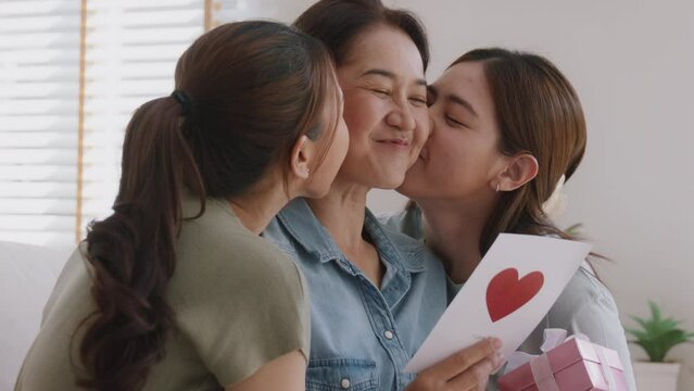 Mother's day two grown up child cuddle hug give flower gift box red heart card to mature mum. Love kiss care mom asia middle age adult three people sitting at home sofa happy smile enjoy family time.