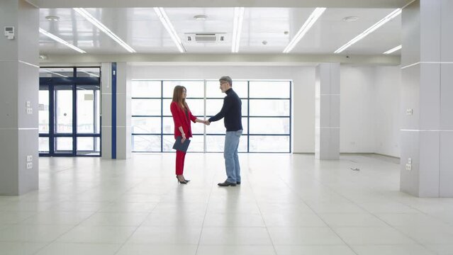 Caucasian grey-haired man shaking hand to woman realtor in red business costume after a conversation about a commercial rental in a white large empty premise