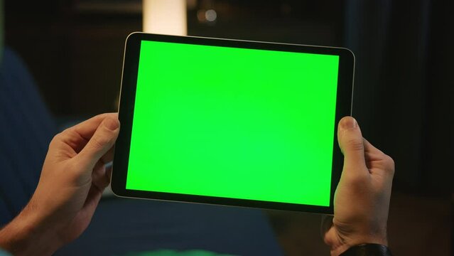 Male hands holding digital tablet with green screen on blurred background of apartment in evening time. Modern technologies, gadget. Indoors