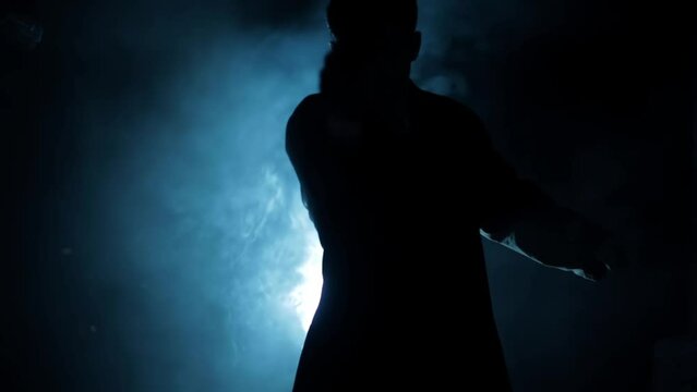 Shot of a man in a long coat and red t-shirt with a pistol in his hand, coming from darkness and shooting. HD footage. Cinematic light with some fog.