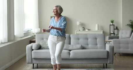 Vivacious older pretty woman moving to favourite music in modern cozy living room. Happy female retiree listen song, dancing at home, spend active life on retirement, enjoy hobby and healthy lifestyle