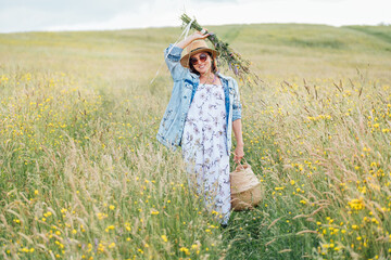 Sincerely smiling young Woman in sunglasses dressed jeans jacket and light summer dress walking by the high green grass meadow with basket and wildflowers bouquet. Human in the nature concept image.