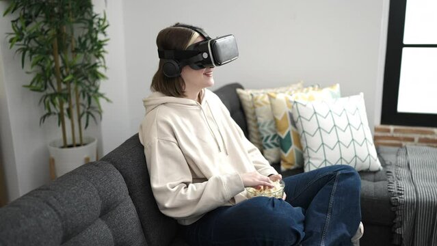 Young blonde woman watching movie using virtual reality glasses eating popcorn at home
