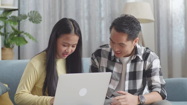 Close Up Of Asian Father Teaching A Daughter Using Laptop While Sitting On A Sofa In The Living Room At Home
