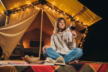 Obraz na płótnie Canvas Smiling Woman freelancer drinking wine and read book sitting in cozy glamping tent in autumn evening. Luxury camping tent for outdoor holiday and vacation. Lifestyle concept