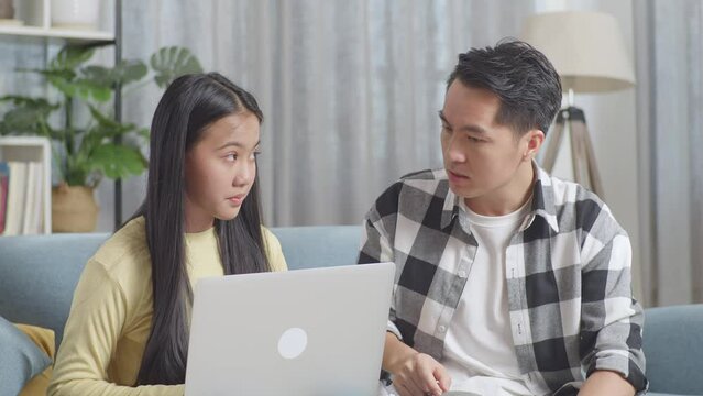 Close Up Of Asian Father Shouting At His Daughter Using A Laptop With Nonsense Things On A Sofa In The Living Room At Home
