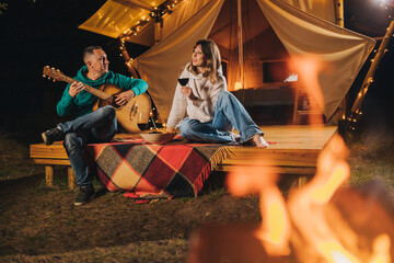Obraz na płótnie Canvas Happy couple relaxing in glamping on autumn evening, drinking wine and playing guitar near cozy bonfire. Luxury camping tent for outdoor recreation and recreation. Lifestyle concept