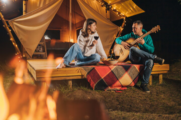 Happy couple relaxing in glamping on autumn evening, drinking wine and playing guitar near cozy bonfire. Luxury camping tent for outdoor recreation and recreation. Lifestyle concept