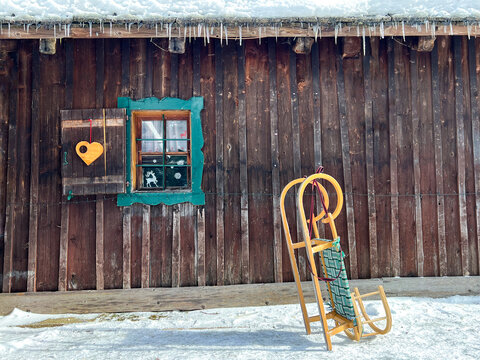 Wooden sleigh stands in the snow against the background of an authentic wooden house with a beautiful window with heart decor  in the high mountains, roof with icicles