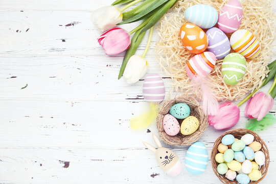 Different colorful eggs in nests and baskets, flowers of tulips on white wooden background