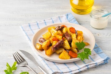 Sliced baked potatoes, carrots and sausages on a ceramic plate on a light wooden background