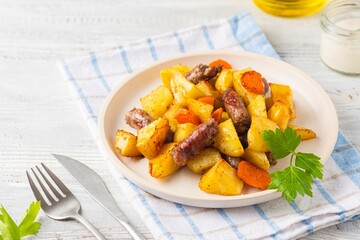 Sliced baked potatoes, carrots and sausages on a ceramic plate on a light wooden background