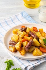 Sliced baked potatoes, carrots and sausages on a ceramic plate on a light wooden background