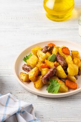 Sliced baked potatoes, carrots and sausages on a ceramic plate on a light wooden background