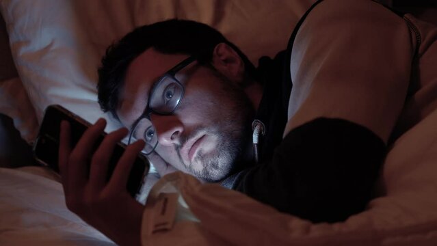 Man lying in bed with smartphone, screen lights up face in dark bedroom, scrolling, social media, texting friends in night before sleep
