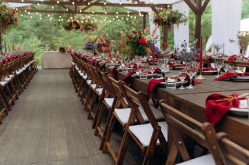 A large, long, wooden table and chairs, with dishes, flowers, candles. Pavilion in nature. Wedding banquet.
