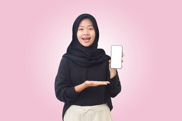 Portrait of excited Asian hijab woman in black outfit showing blank screen mobile phone and pointing on it with finger. Advertising concept. Isolated image on pink background