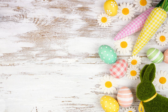 Easter side border with pastel eggs, farmhouse style cloth carrots, flowers and moss bunny. Above view over a white wood background. Copy space.