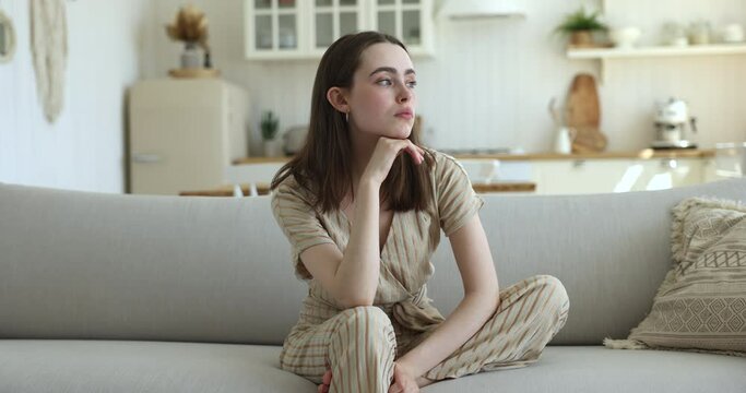 Beautiful young 20s woman reflecting seated on sofa, feels bored on weekend alone at home, looks thoughtful deep in sad thoughts, waiting or missing for boyfriend, suffers from loneliness, recollects