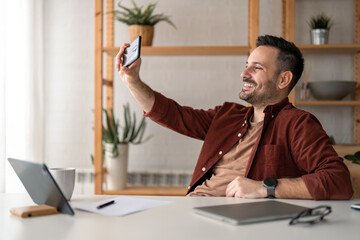 Smiling handsome male freelance worker using smartphone, looking at front camera, taking a selfie...