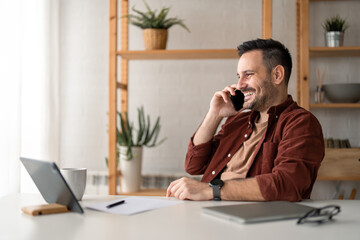 Young handsome businessman using modern smartphone, having a call in home office, sitting at desk,...