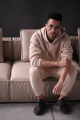 Portrait of a cheerful young african american man 20s wearing casual hoody and glasses sitting on sofa against black background. copy space isolated