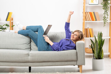 Fototapeta premium Portrait of an enthusiastic girl in full length, lying on the sofa, raising her fist, with a phone and a laptop