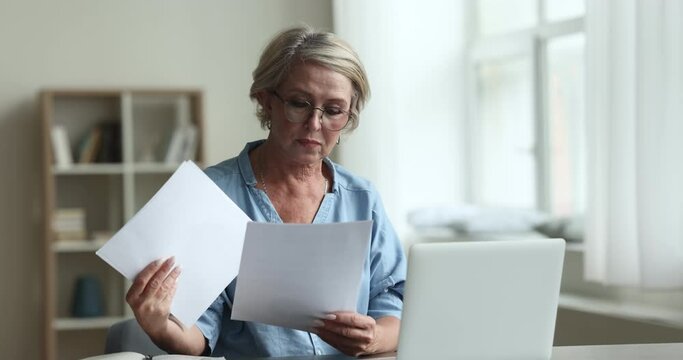 Mature woman in glasses sits at desk with laptop and sorting out bills, papers, learn received legal documents, reviewing pension status, family budget management, make payments through e-bank system