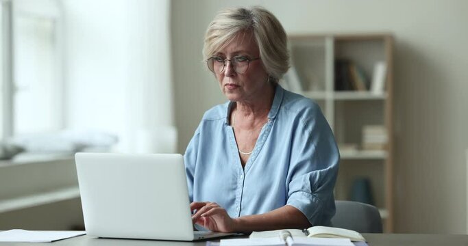 Focused aged woman in glasses doing telework on retirement on laptop. Serious mature female working, search information on internet, ponders looks concentrated, thoughtful engaged in workflow on-line