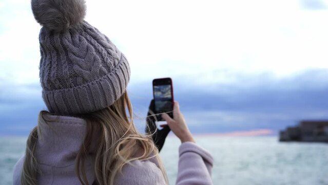 Girl in a hat and coat takes pictures of the sea coast with a smartphone
