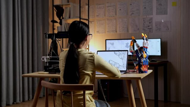 Back View Of Teen Asian Girl With 3D Printing Stretching While Designing A Cyborg Hand On A Laptop At Home
