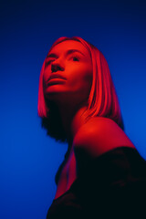 Young female with fair hair looking at camera against blue backdrop in studio with neon illumination