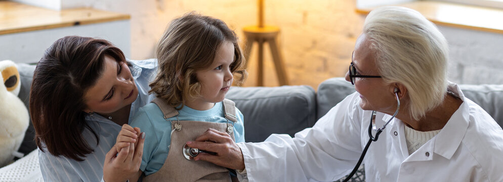 Kind female senior paediatrician doctor visiting his patient at home, examining little girl sitting on mother's lap, writing prescription. Concept of kid's health check. Successful recovery. Banner - Powered by Adobe