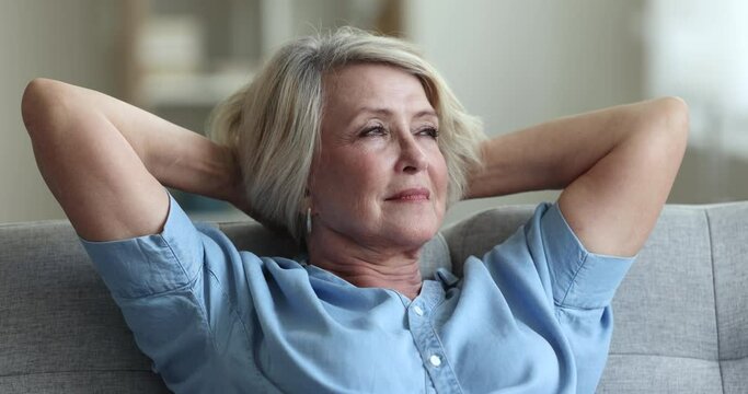Carefree senior pretty woman smile, relax on cozy sofa, take break, breath fresh conditioned air inside warm living room, put hands behind head enjoy untroubled retired life, close up. Daydreams, rest