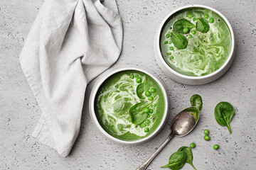 Two ceramic bowls with spinach cream soup with green pea and fresh spinach leaves on grey concrete...