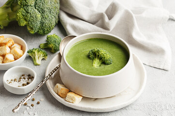 Green vegetable cream soup with broccoli and croutons in ceramic bowl over gray concrete background with ingredients. Side view
