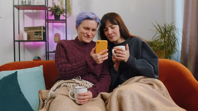 Two young and cheerful lesbian or friends hold their smartphones while shopping on e-commerce apps at home living room. Girls enjoy having fun browsing different social media applications together