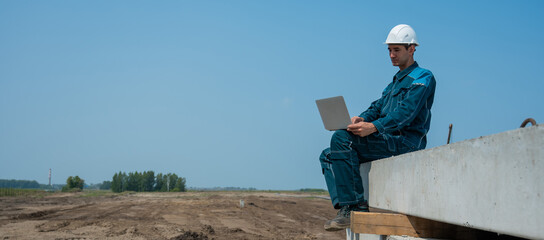 Caucasian male builder in hardhat sits on floor slabs and uses laptop at construction site. 